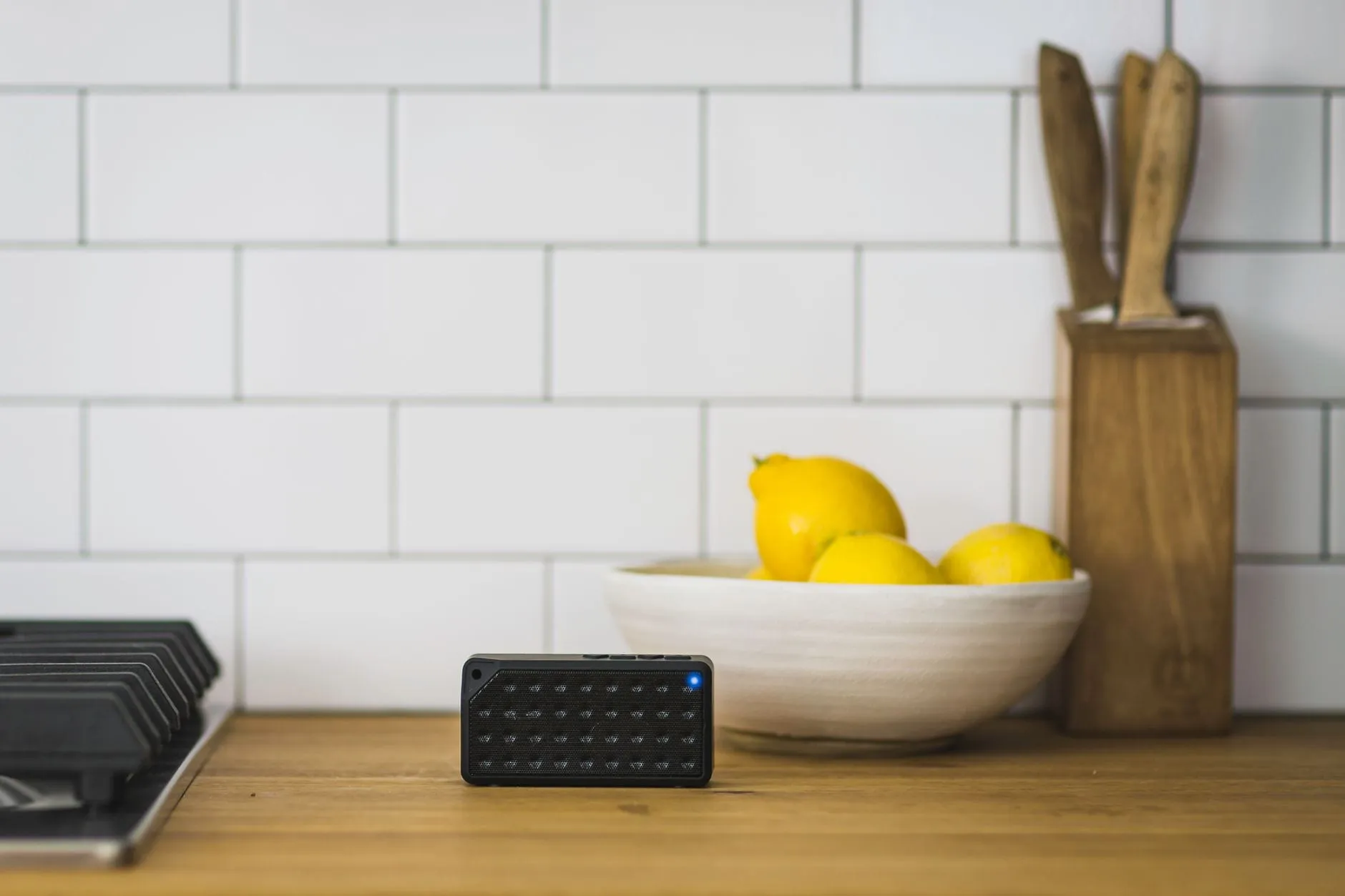 A small black wireless speaker with its status light lit, standing on a wooden kitchen counter in front of white tiles beside a bowl of lemons.