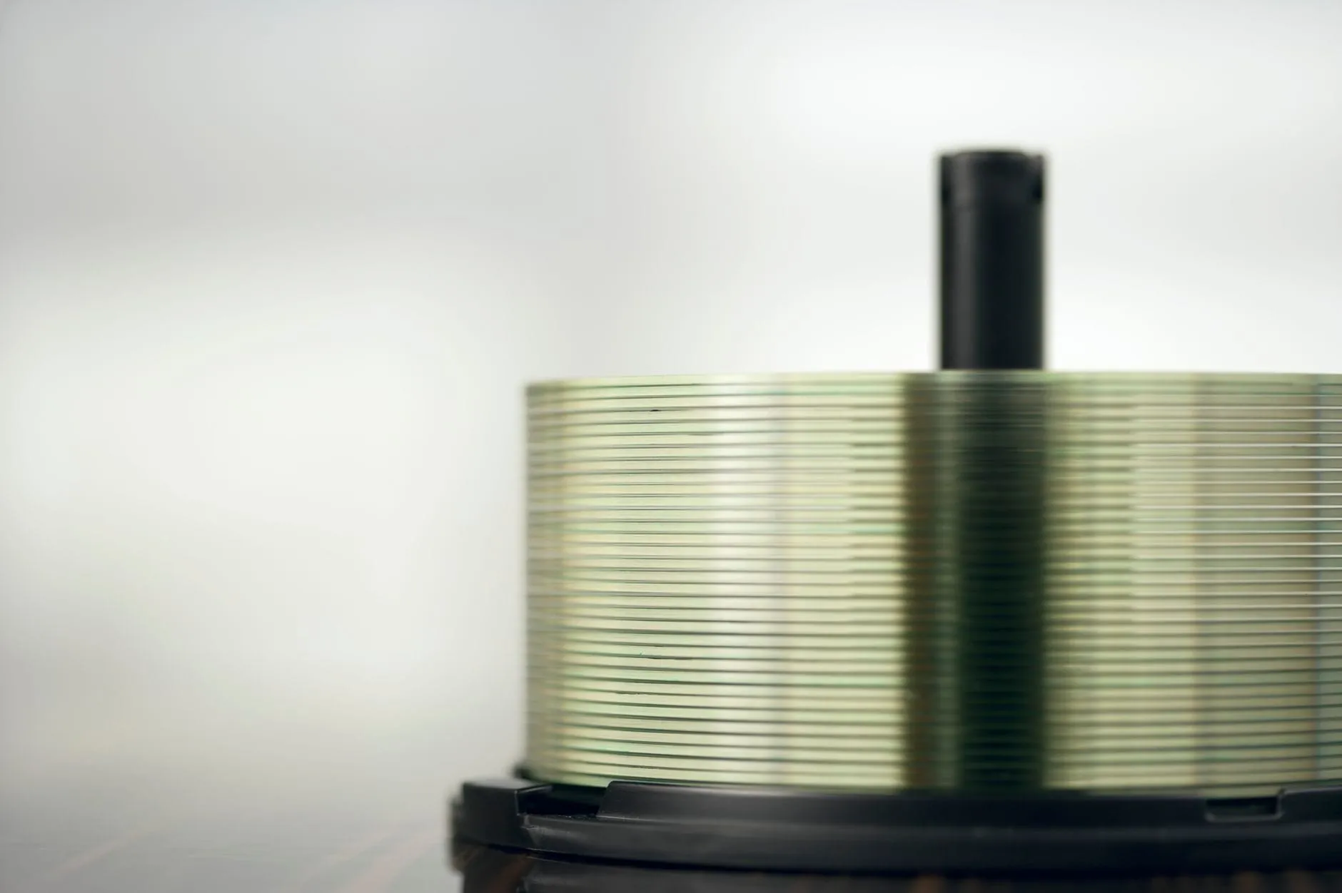A tall stack of unlabelled optical discs on a black spindle, their edges catching a faint green sheen against a pale background.