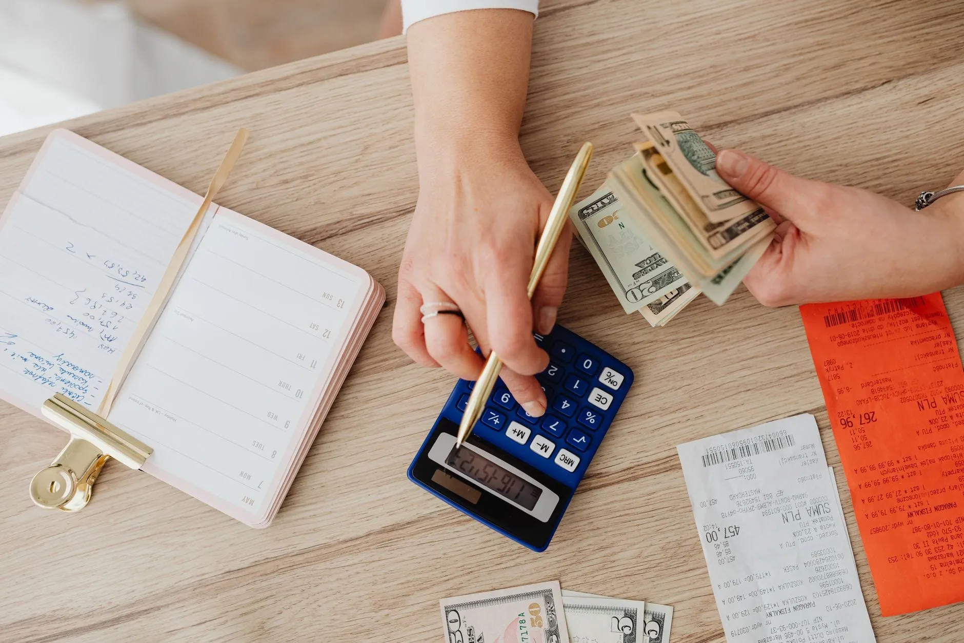 Hands working a pocket calculator on a wooden table strewn with till receipts, banknotes and a notebook of handwritten sums.