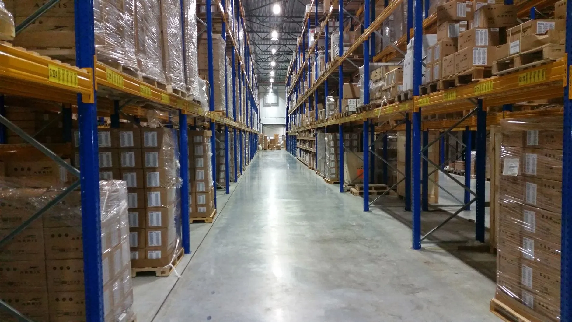 A warehouse aisle between tall racks of palletised boxes, each bay carrying a yellow location label such as G224.