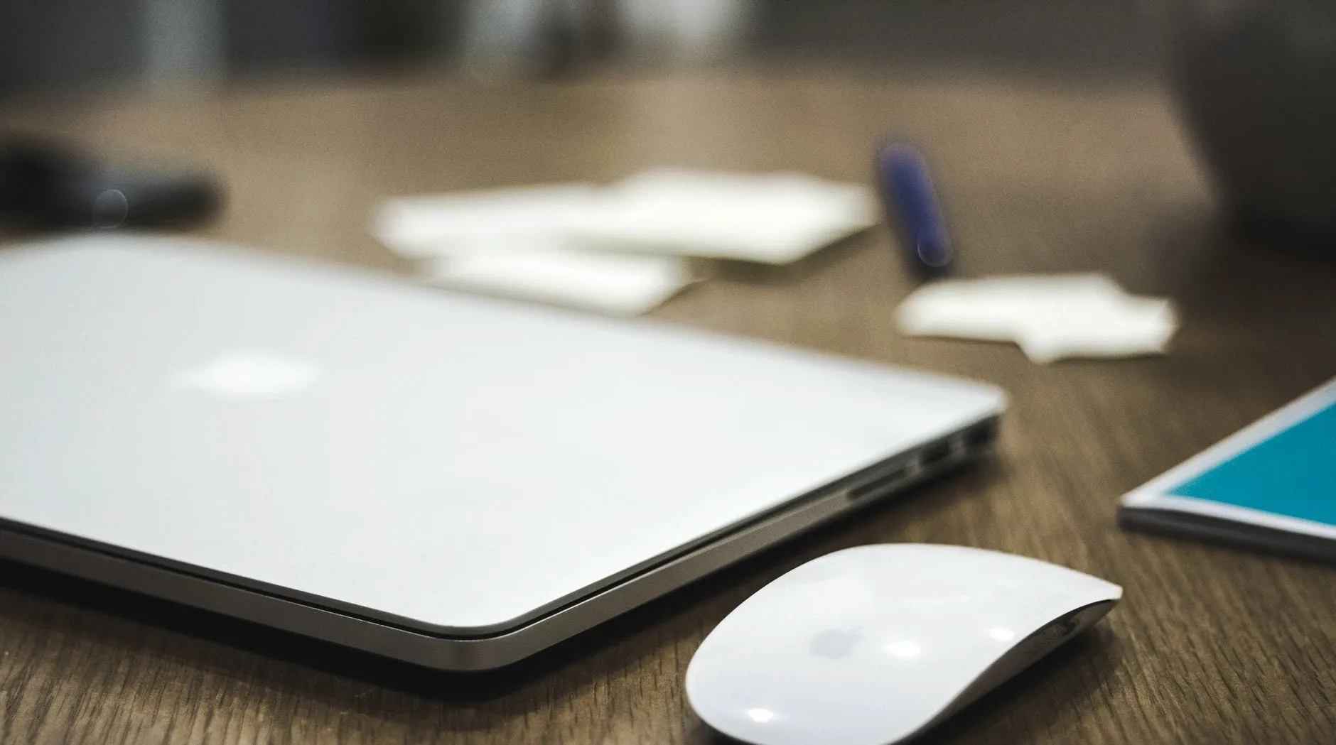 A closed silver laptop and a white mouse on a wooden desk, with sticky notes and a pen just out of focus behind them.