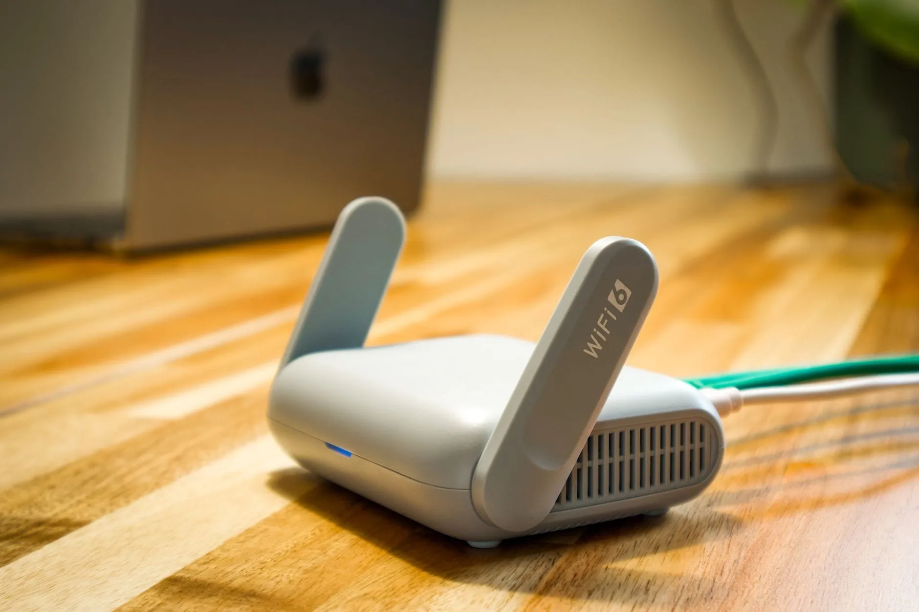 A small white router with two raised antennas on a wooden floor, a green ethernet cable plugged into the back and one antenna marked WiFi 6.