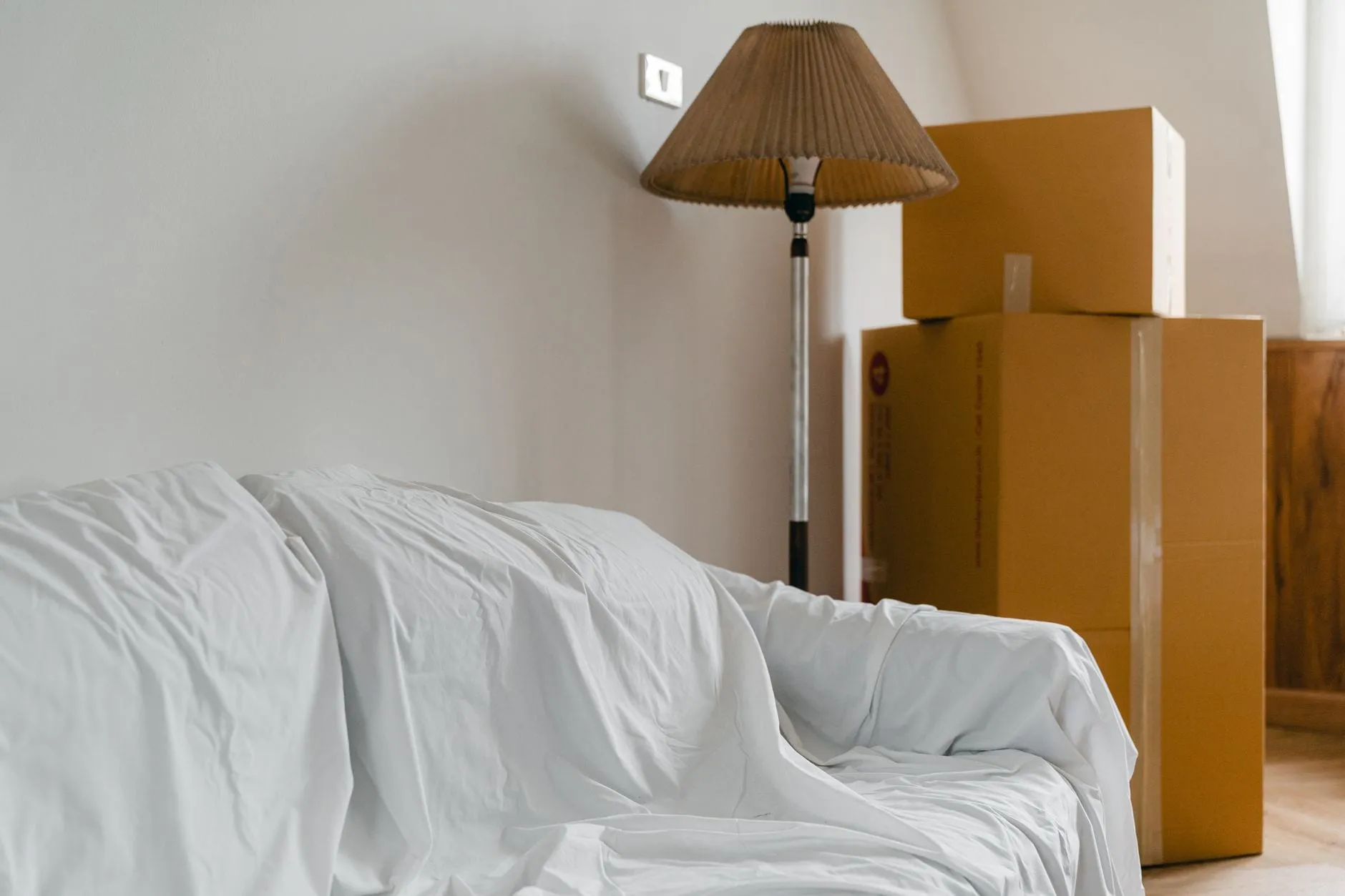 A room part-way through a move: a sofa hidden under a white dust sheet, a floor lamp, and sealed cardboard boxes stacked in the corner.