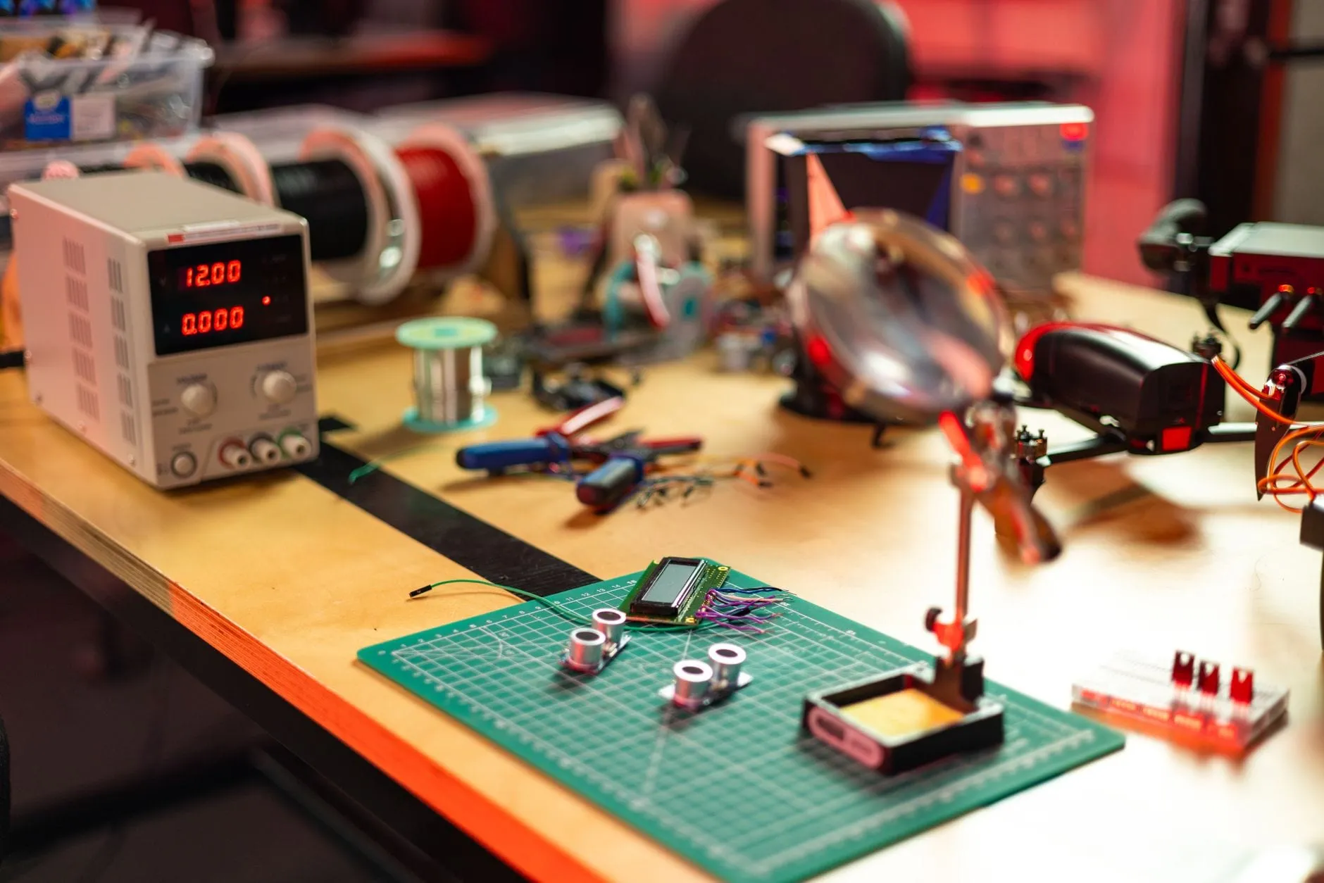 An electronics workbench with a bench power supply reading 12.00 volts, a spool of solder, jumper wires and small sensor modules laid out on a green cutting mat.