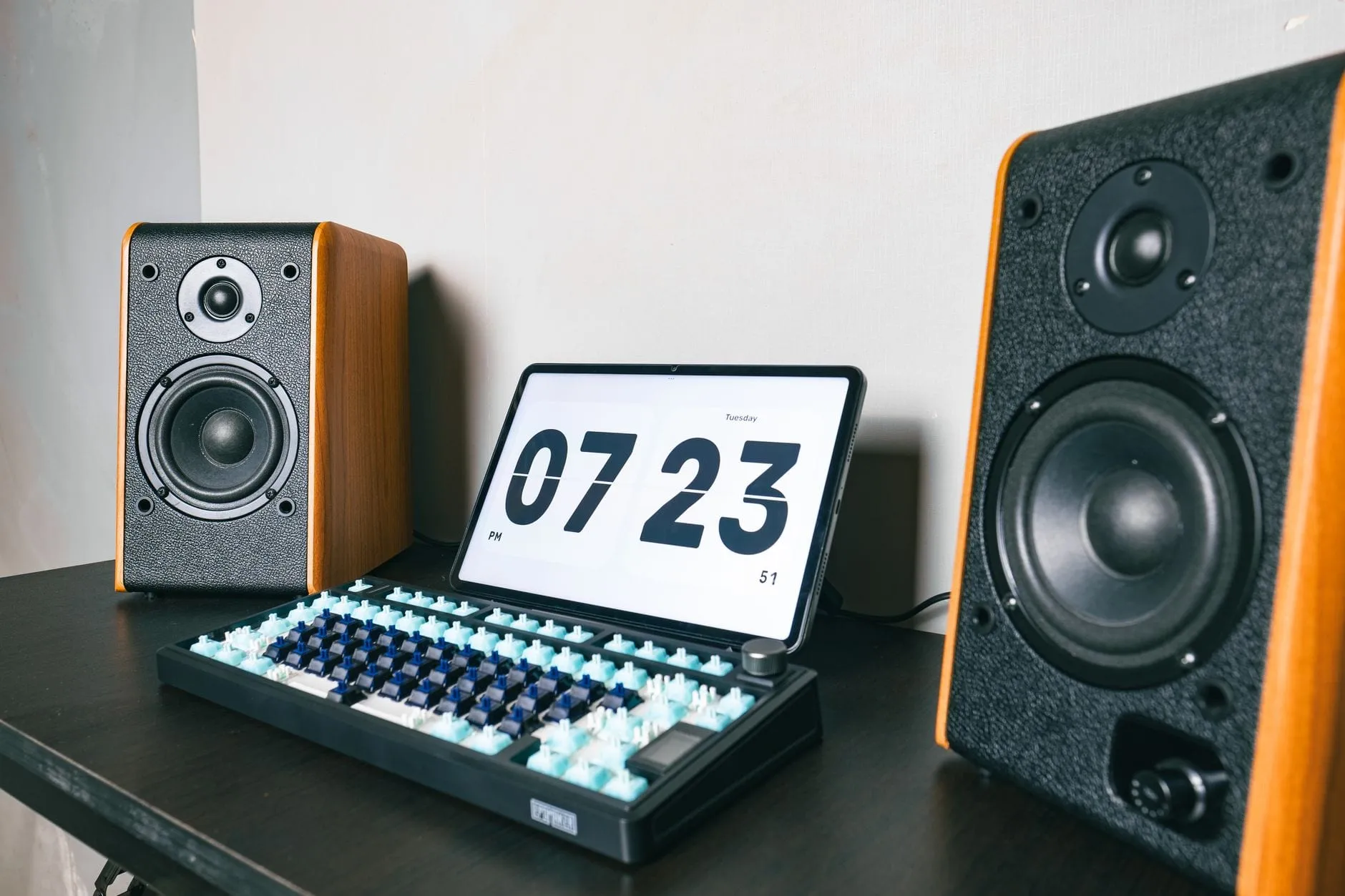 A pair of wood-sided bookshelf speakers either side of a tablet showing a flip clock, with a partly built mechanical keyboard on the desk between them.