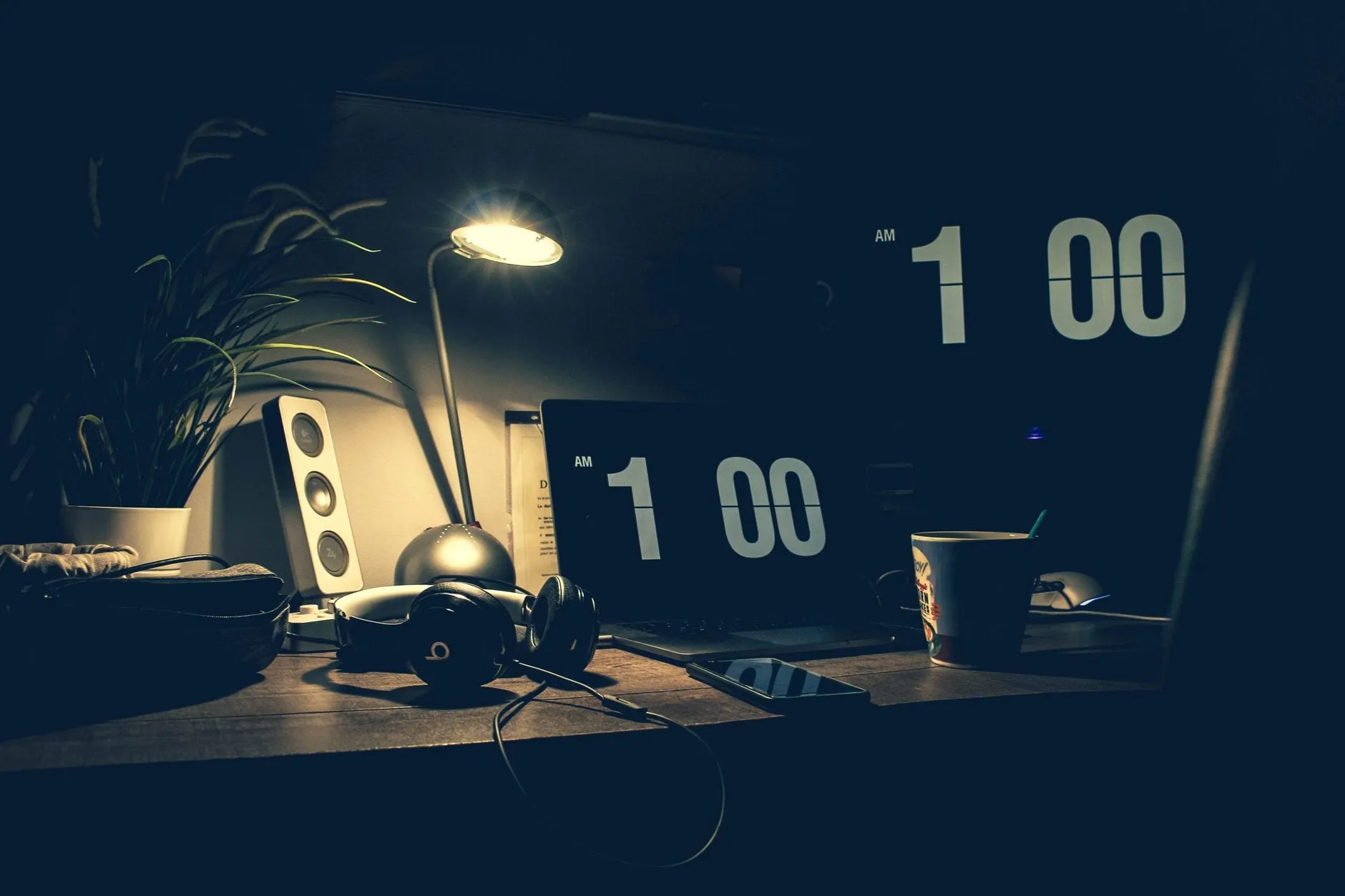 A desk at night lit by a single lamp, with a laptop showing a flip clock reading 1:00, wired headphones, a small speaker and a mug.