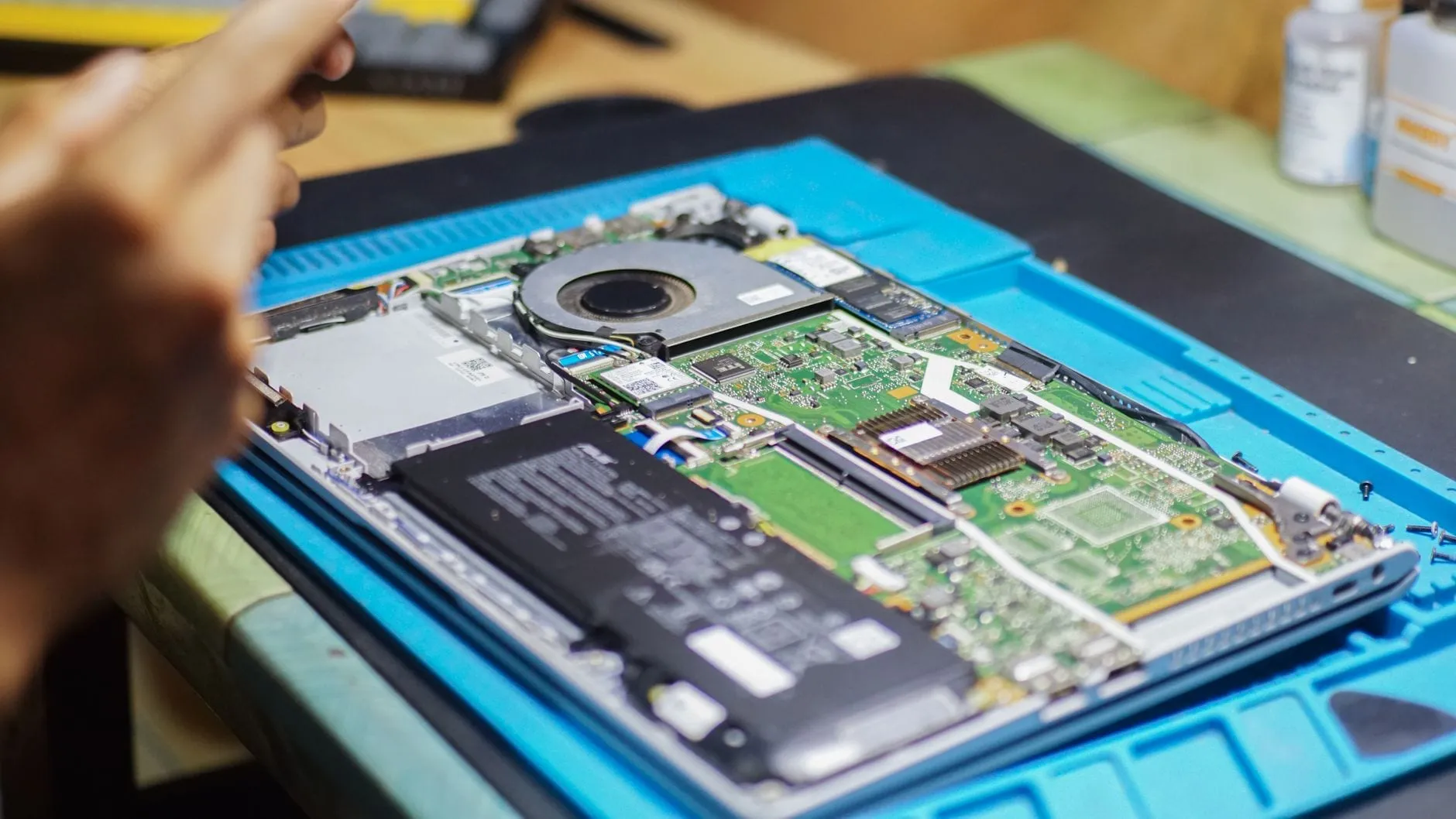 A laptop opened on a blue repair mat with its battery, cooling fan, mainboard and ribbon cables exposed.