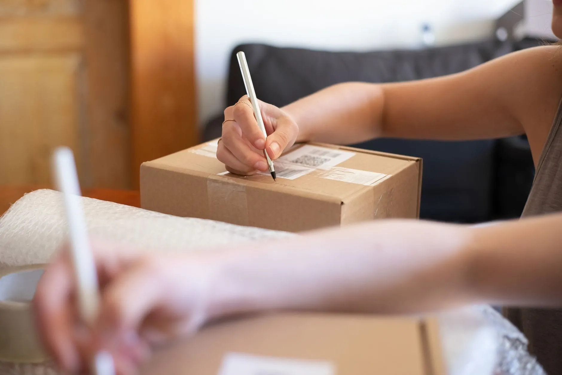 A hand writing on the label of a sealed cardboard box while a second pair of hands works with bubble wrap alongside.