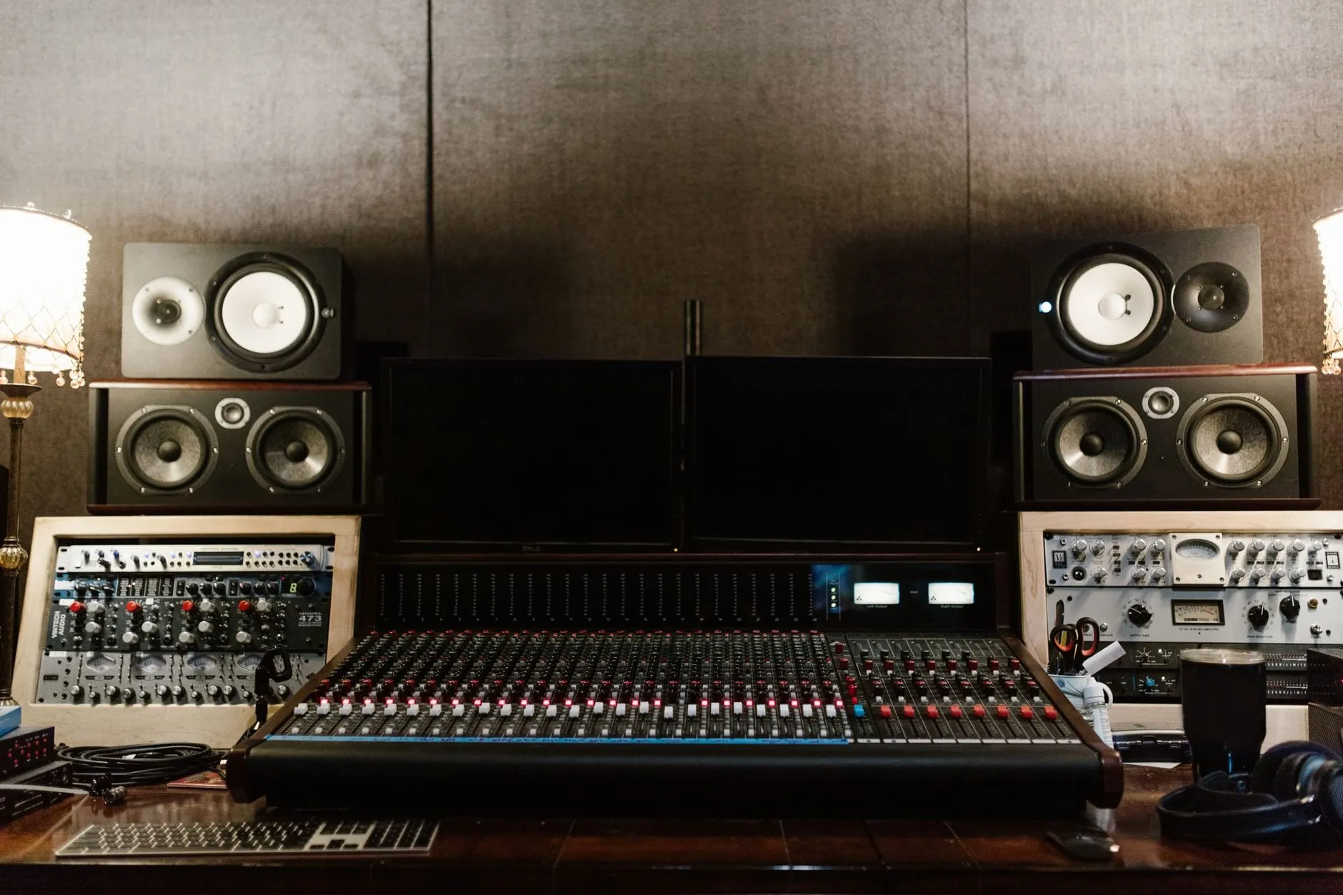 A studio control room: a large mixing console with rows of faders, two pairs of monitor speakers on either side, and outboard racks flanking dark screens.