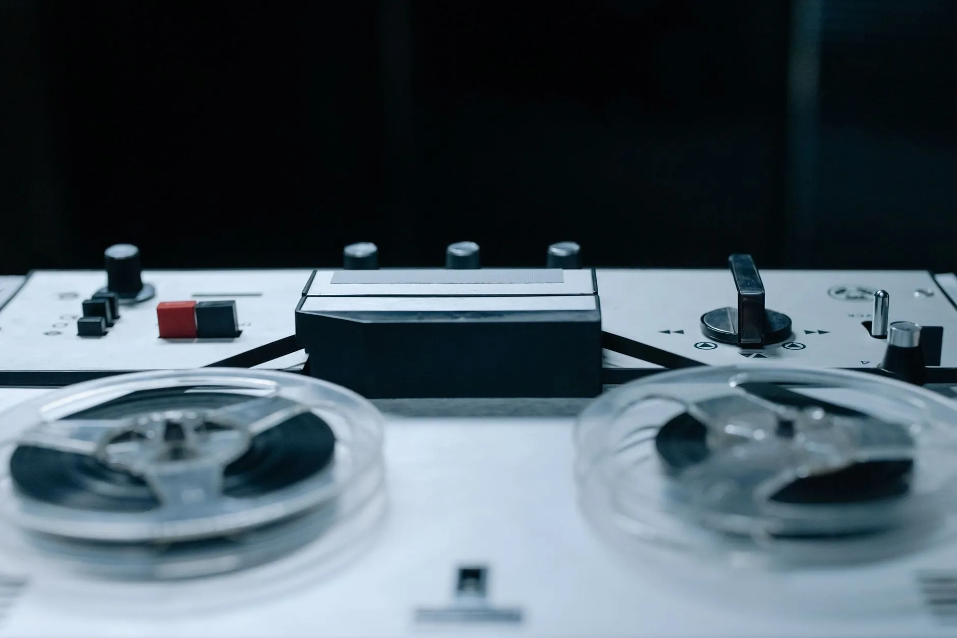A reel-to-reel tape machine seen close up, two clear spools of tape mounted either side of its head cover and transport controls.