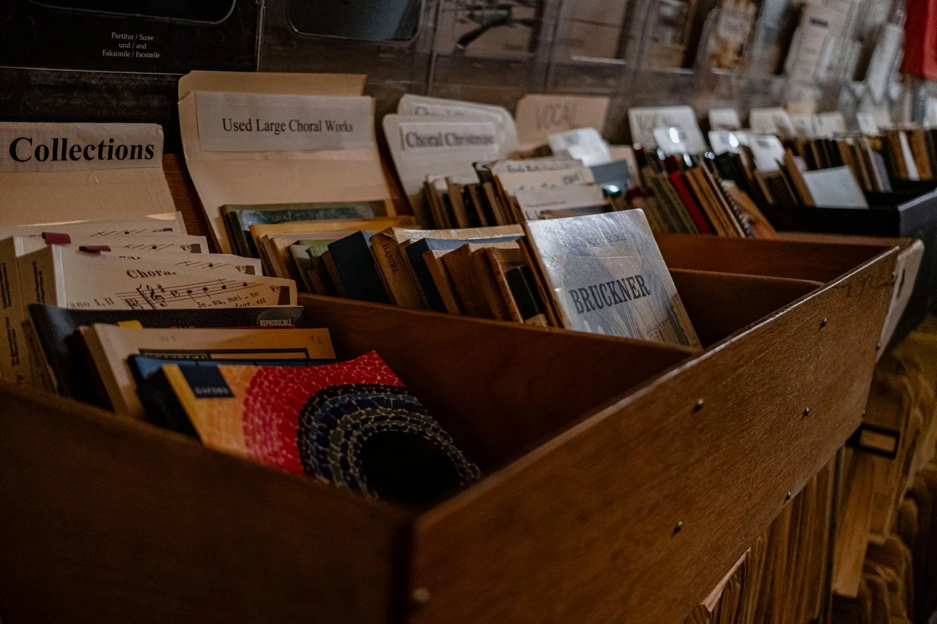 Sheet music filed upright in wooden bins behind printed dividers reading Collections, Used Large Choral Works and Choral Christmas, with composer scores visible between them.