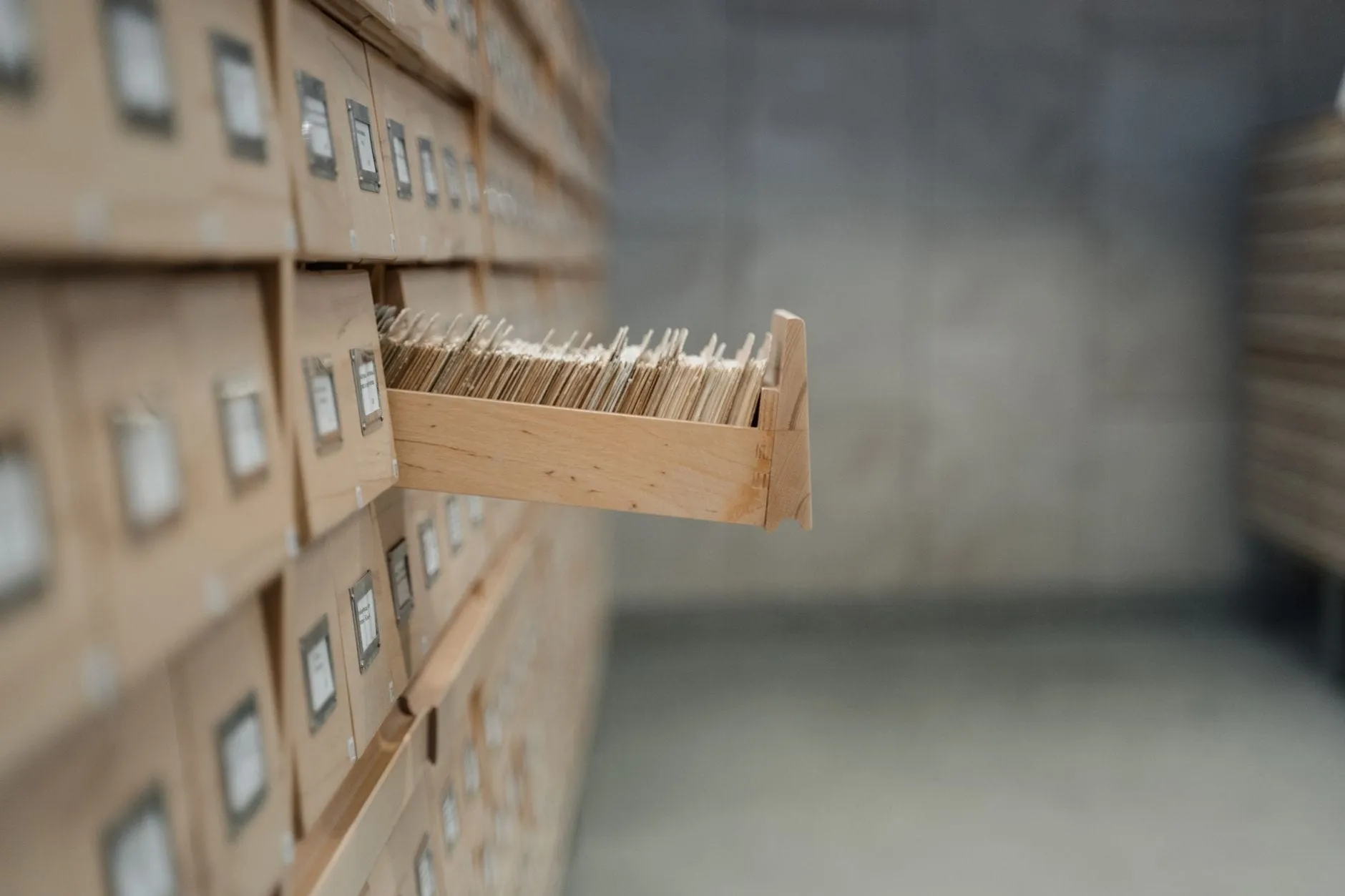 A wall of pale wooden card-catalogue drawers in an archive, one drawer pulled open to show it packed tight with index cards.