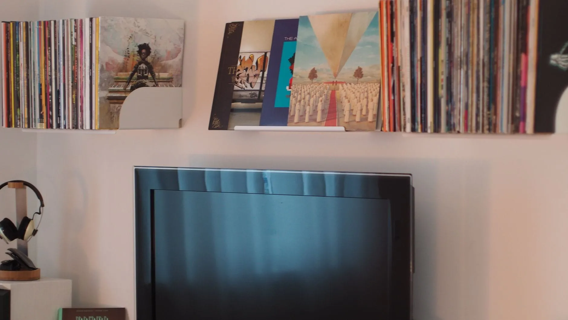 A wall shelf packed with vinyl records above a wall-mounted television, with headphones resting on a wooden stand to one side.