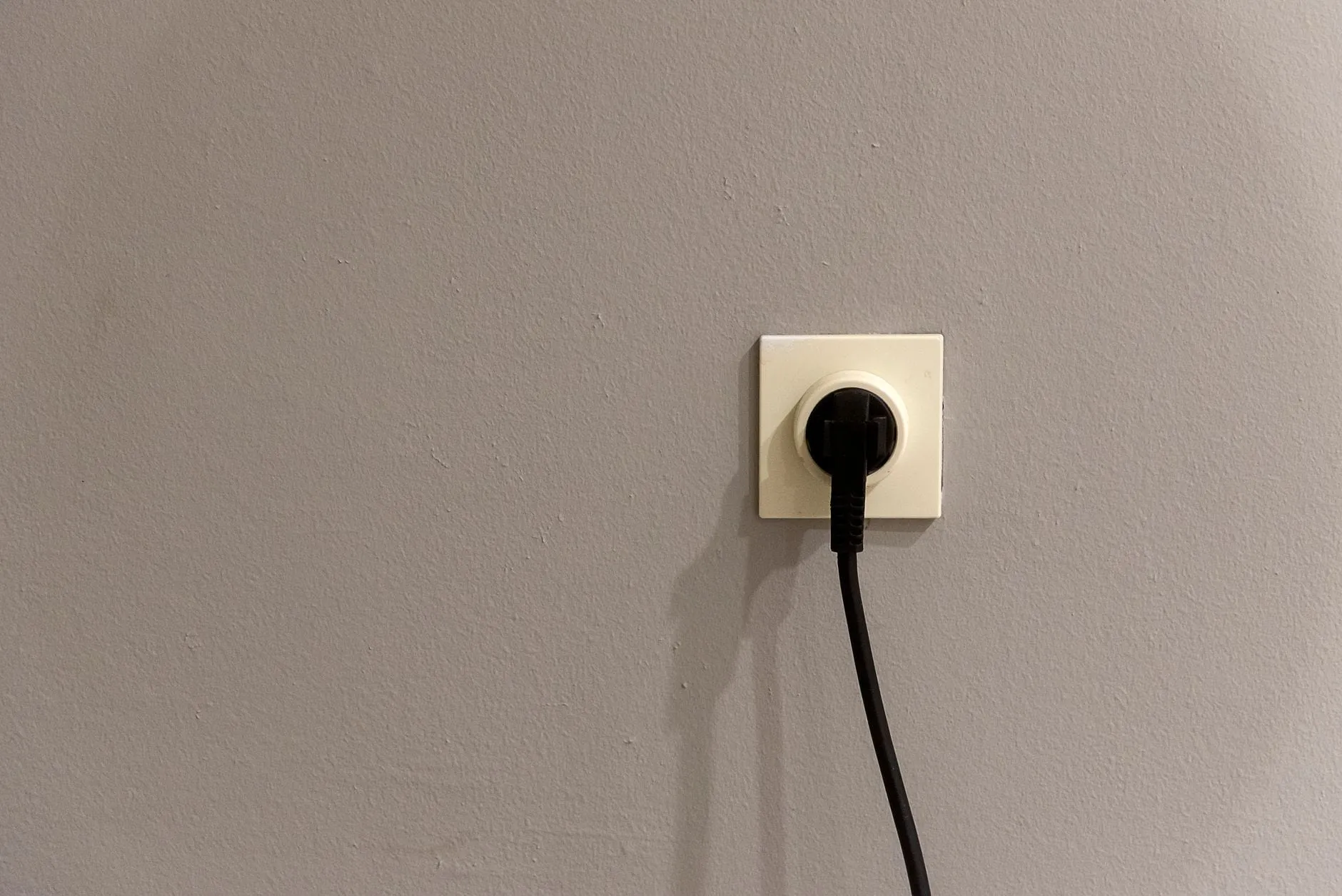 A single black plug seated in a cream wall socket on a plain painted wall, its cable trailing down out of frame.