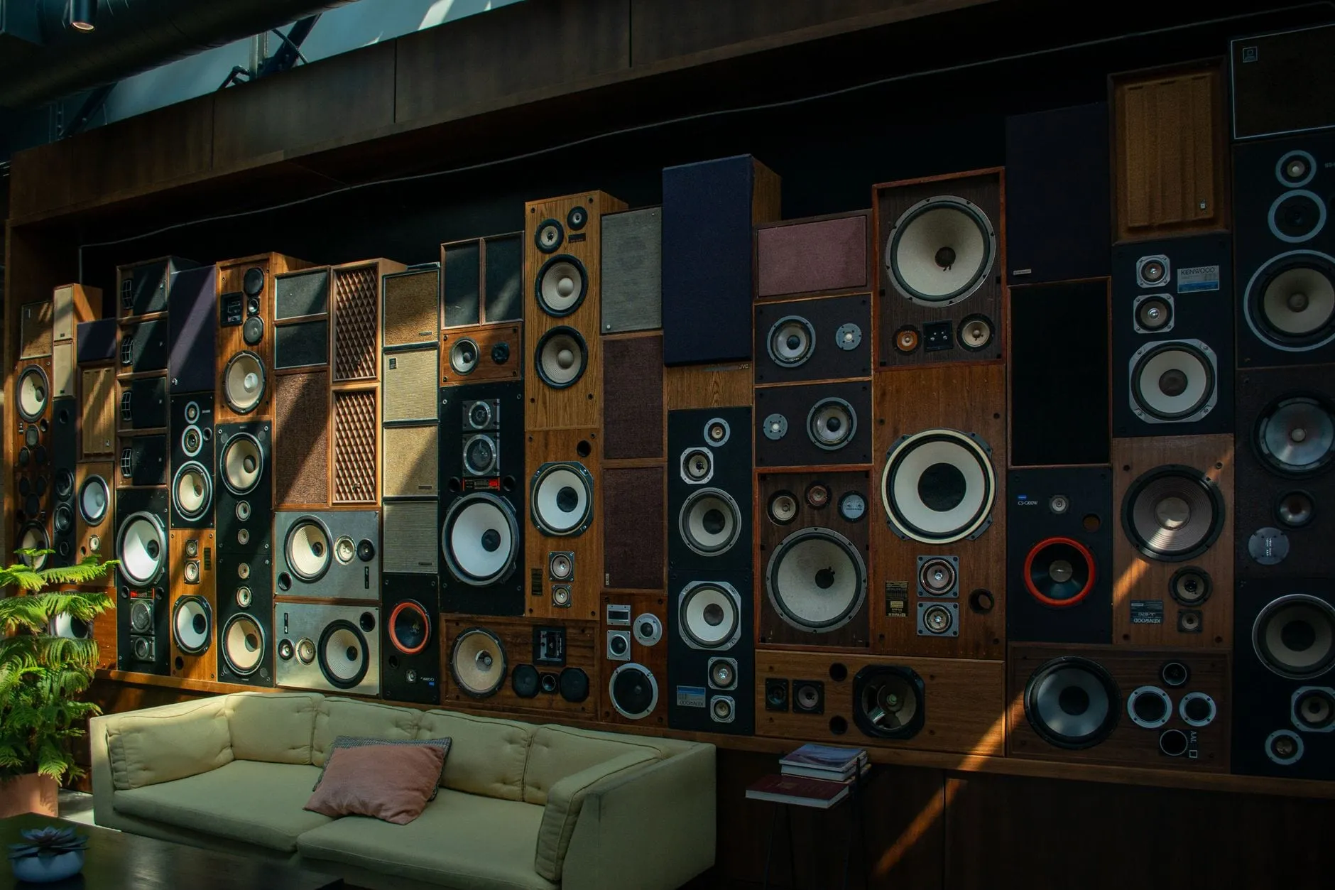 A wall stacked floor to ceiling with dozens of mismatched vintage loudspeaker cabinets of different sizes and finishes, above a pale sofa.