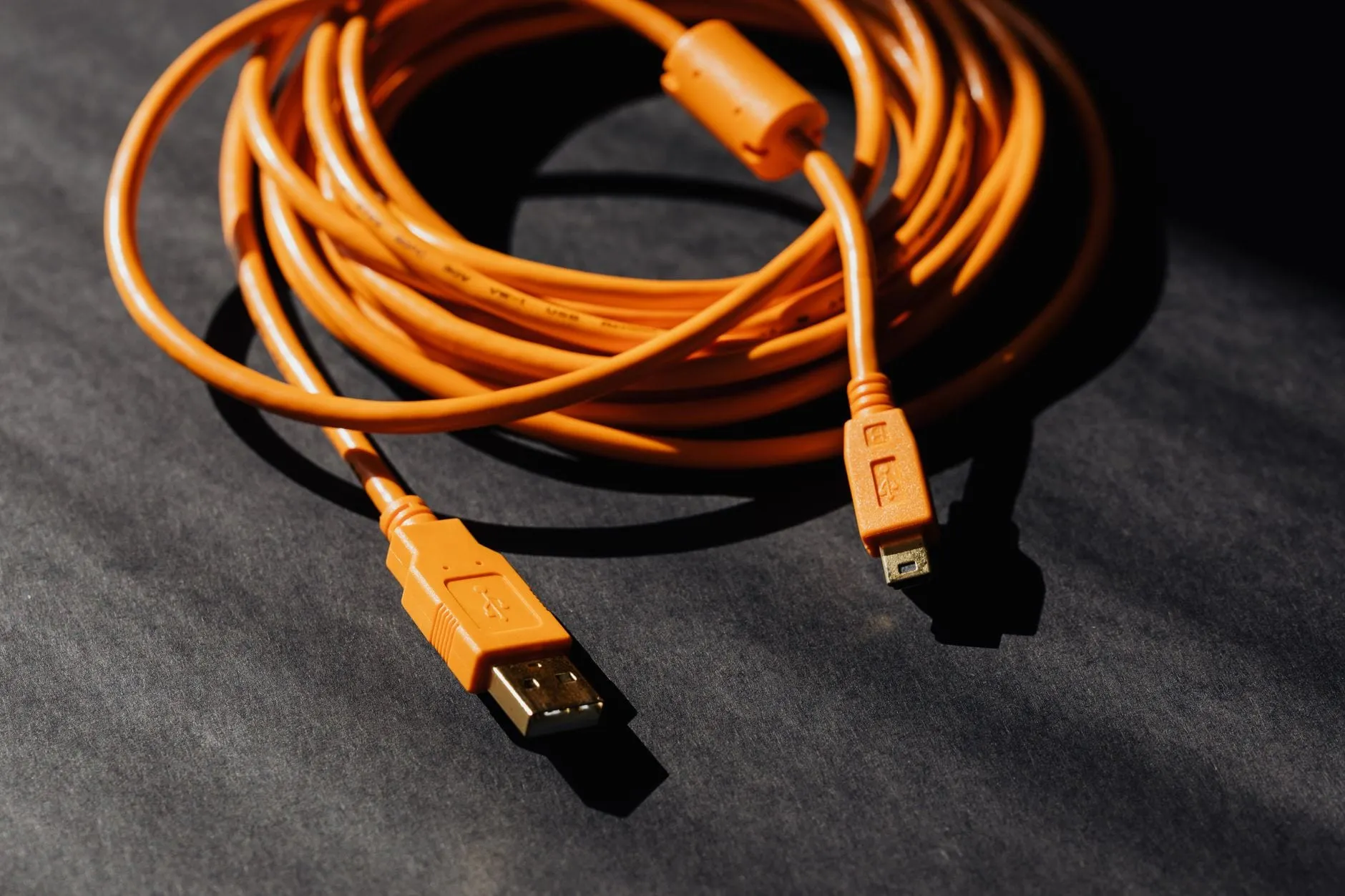 A coiled orange USB cable on a dark surface with both connectors laid out in front of it and a ferrite bead near one end.