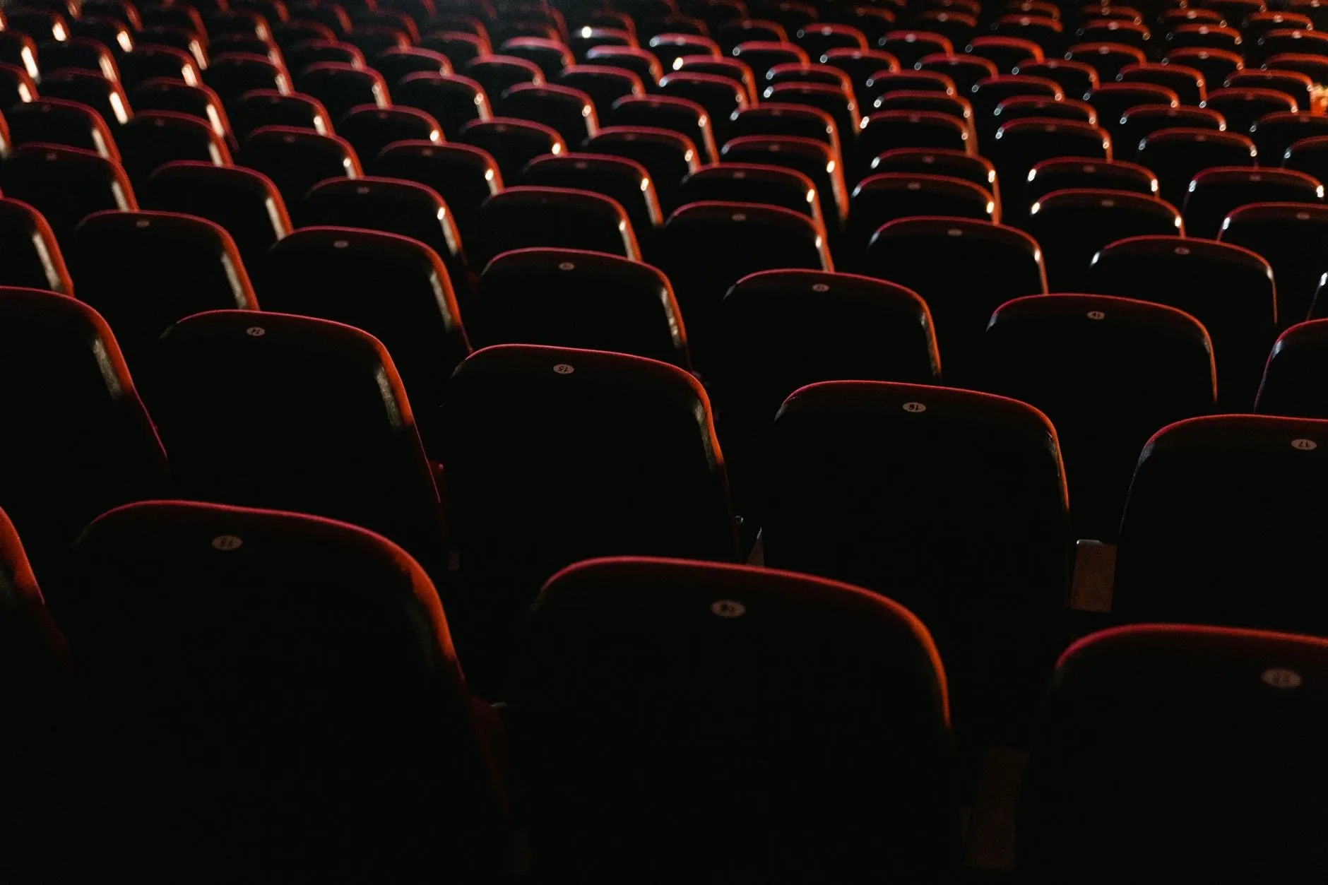 Rows of empty red cinema seats in low light, each seat back carrying a small numbered disc.