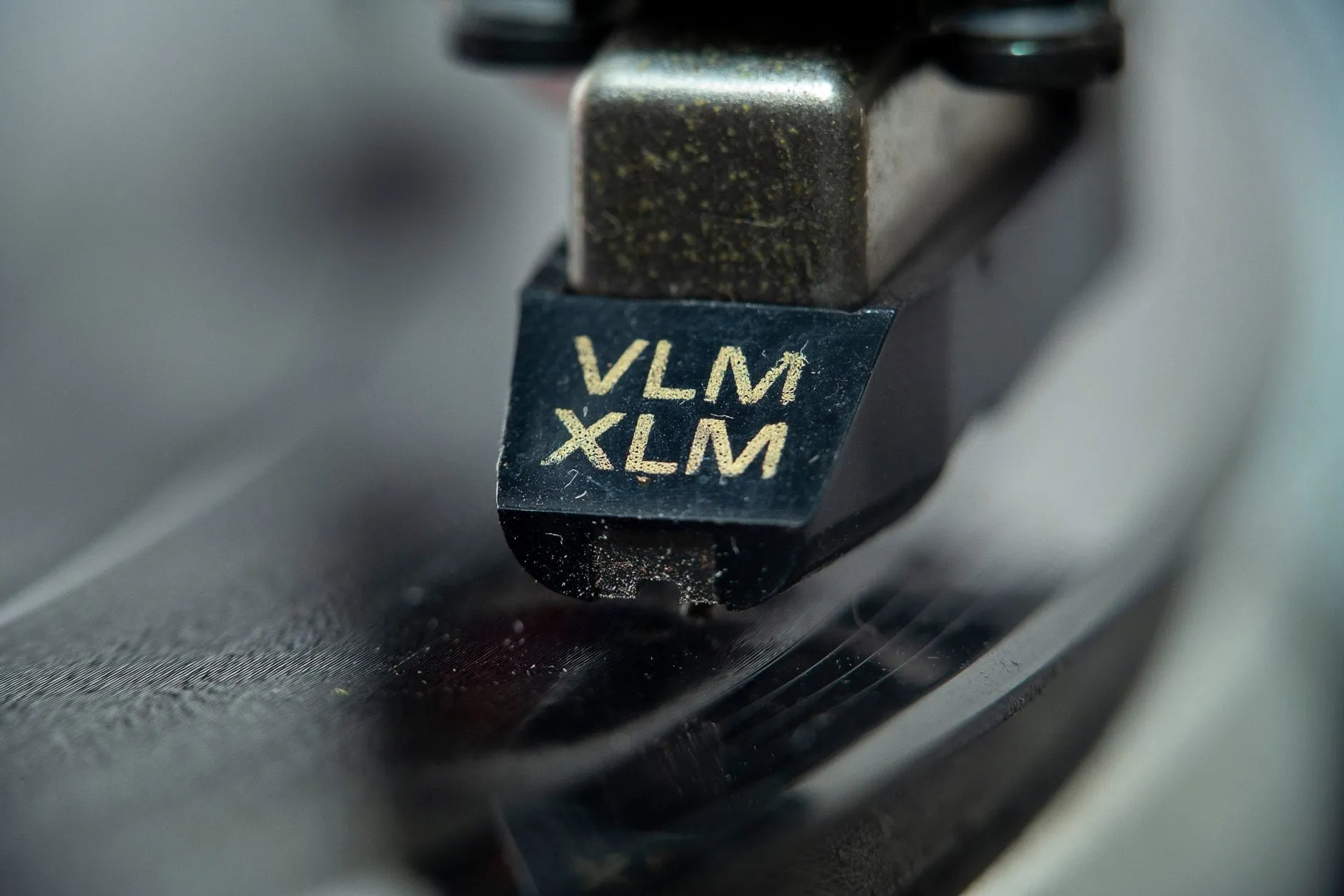 A phono cartridge photographed in extreme close-up, its stylus down in the groove of a spinning record, the body marked VLM XLM.
