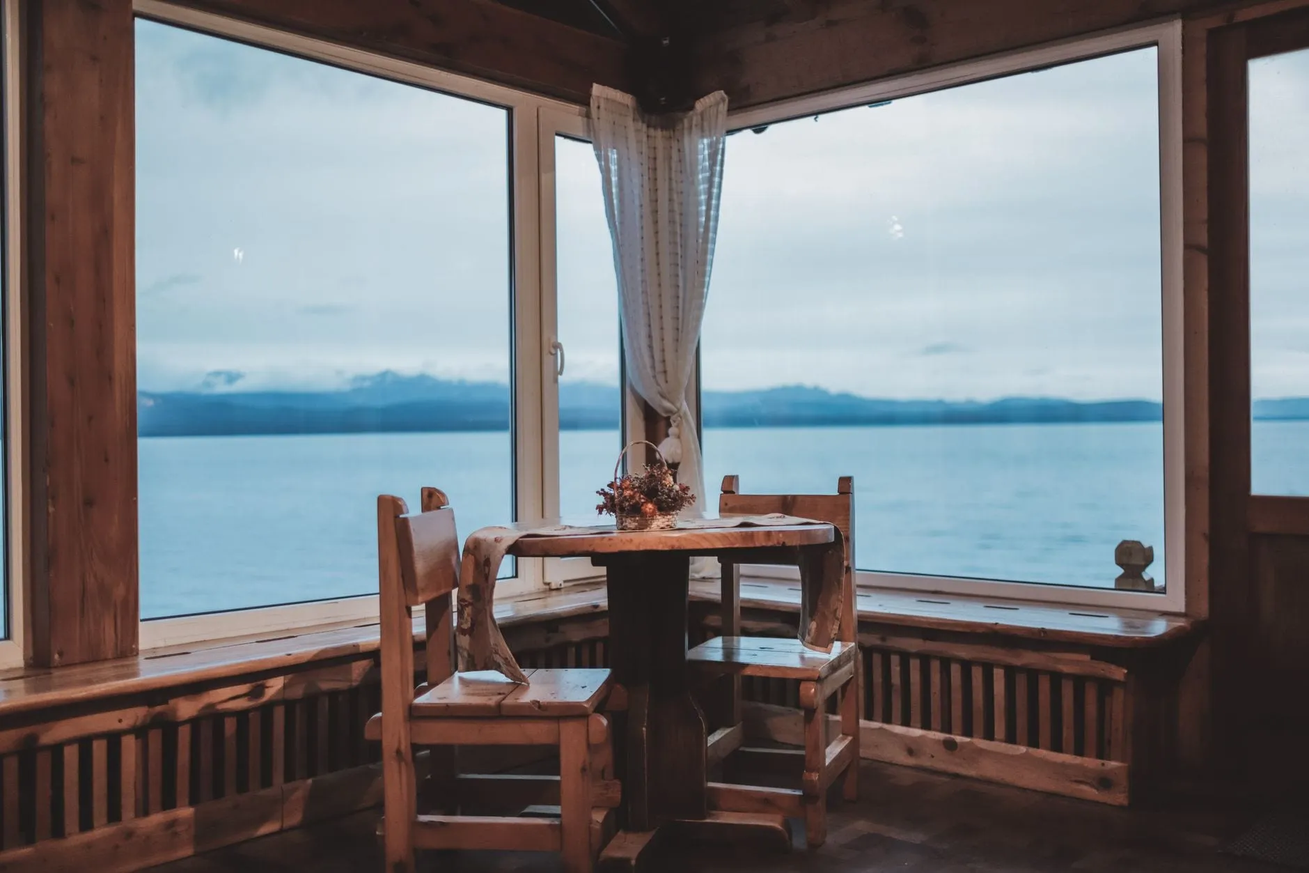A corner room panelled in timber, a small table and two chairs at the window, with a wide lake and distant mountains beyond the glass.